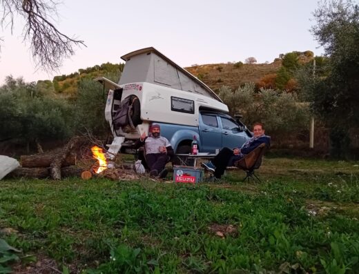 Two people sitting around a campfire in front of a camper van.