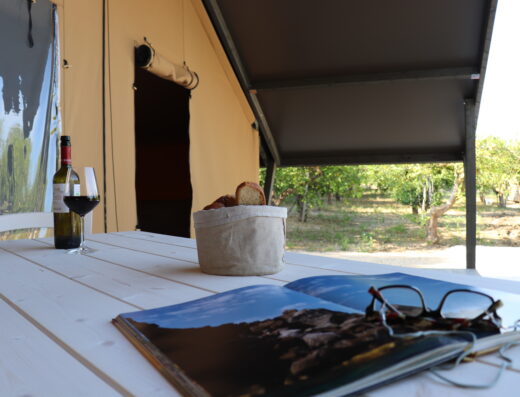 A picnic table set with a bottle of wine, a basket of bread, and an open magazine, with a canvas tent in the background.