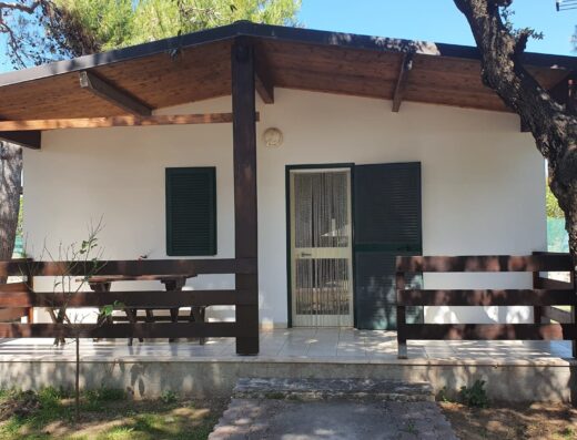 A single-story house with green shutters, a small porch, and a picnic table set under the shade of trees.