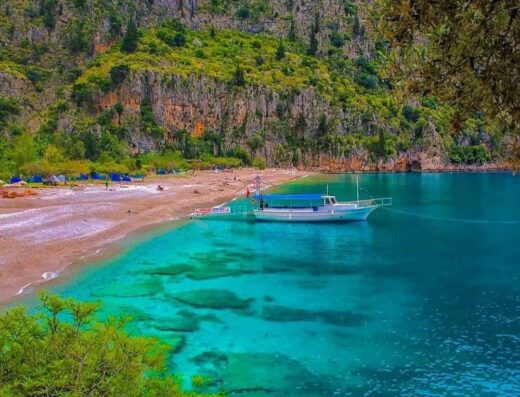 A tranquil cove with clear turquoise waters, a pebbly beach, and a single boat moored near the shore, all surrounded by lush greenery and steep cliffs.