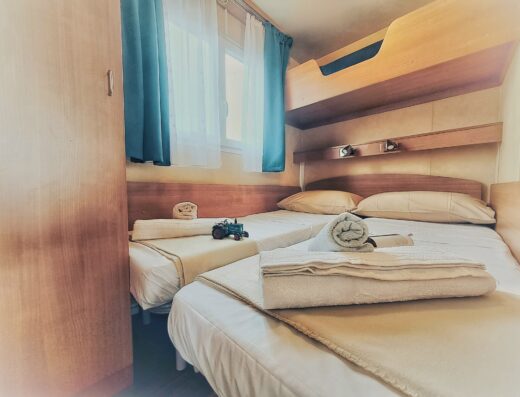 Interior of a cozy bedroom with two beds, one bunk bed above a double bed, towels folded on each and sunlight filtering through blue curtains.