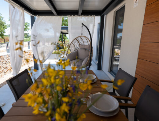 A cozy outdoor dining area with a set table, flowers, and a hanging chair on a sunny day.