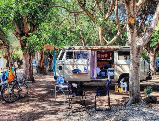 Camper van parked under the shade of trees with chairs and a bicycle nearby at a campsite.