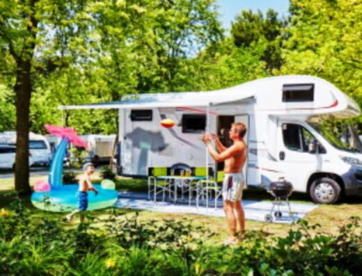 A family enjoys outdoor activities near their motorhome in a sunny campground with lush green trees.
