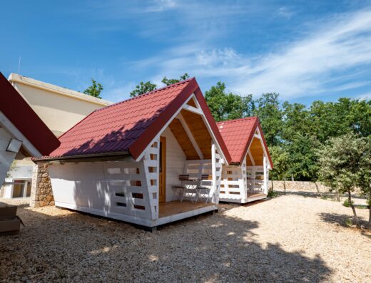 Two small white cabins with red roofs in a gravel outdoor area near trees.