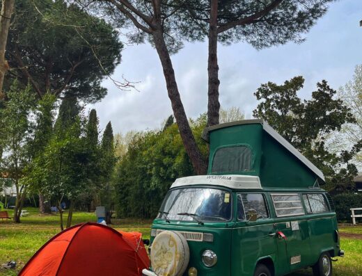 Vintage green camper van with a pop-up roof parked next to a red tent under tall pine trees in a camping area.