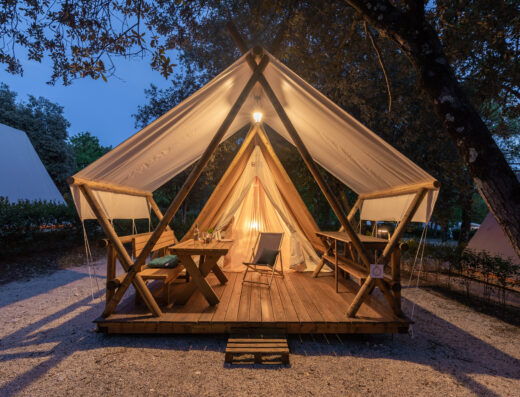 Luxurious glamping tent with open flaps, interior lights on, set on a wooden platform in a twilight forest setting.