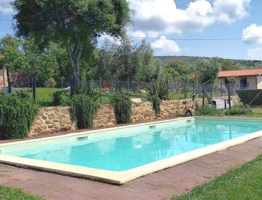 A rectangular swimming pool surrounded by a lush green garden and stone walls under a clear blue sky.