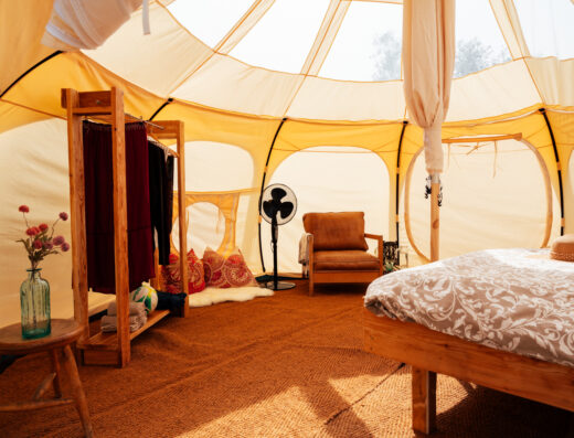 Interior of a luxurious glamping tent with a comfortable bed, armchair, and wooden furniture, bathed in warm, natural light.