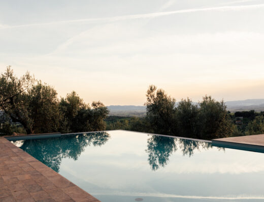 An infinity pool reflecting the sky is surrounded by a stone deck with trees and hills in the background at sunset.