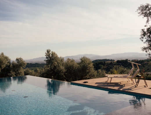 An infinity pool overlooking a scenic landscape with mountains in the distance, surrounded by trees. Two lounge chairs and a small table are placed on the poolside patio.