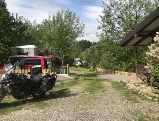 Two parked motorcycles and a red camper van with a rooftop tent along a gravel path, surrounded by trees and greenery, with a small wooden fence on the right.