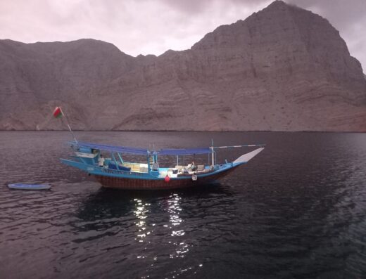 A traditional blue boat with a small red and green flag floating on calm water near a large rocky mountain under an overcast sky.