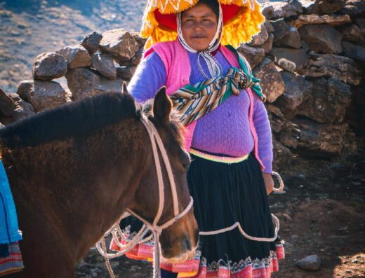 A person wearing colorful traditional clothing stands next to a brown horse in a rocky mountainous area.
