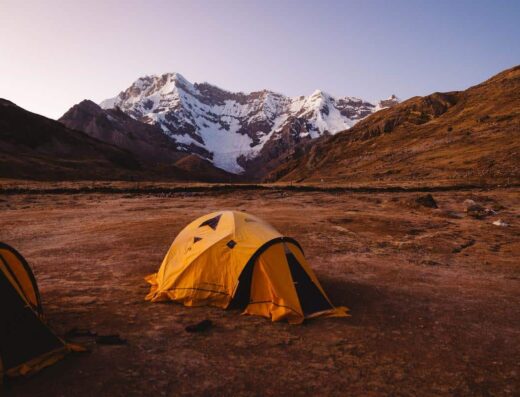Two yellow tents set up on barren ground with snow-capped mountains in the background under a clear sky at dusk.
