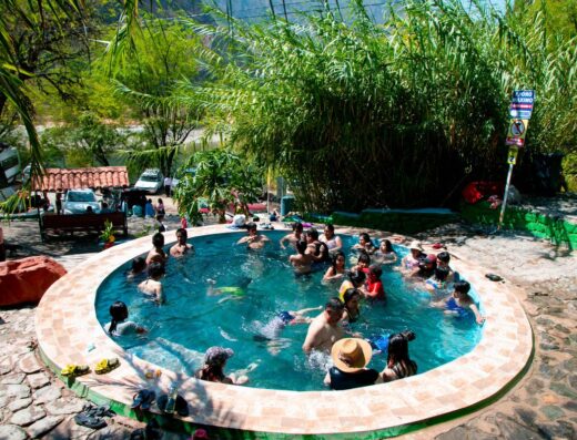 A group of people swim and relax in a round outdoor pool surrounded by lush greenery and stone paving. Nearby, some individuals sit on benches and a few vehicles are parked at the edge of the scene.