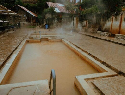 An outdoor thermal pool with murky water, surrounded by stone walls and buildings, with steam rising from the water. The area appears quiet with no people present.