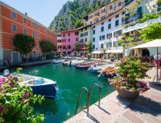A small harbor with several boats docked is surrounded by colorful buildings and lush greenery, with a steep rocky hill in the background.