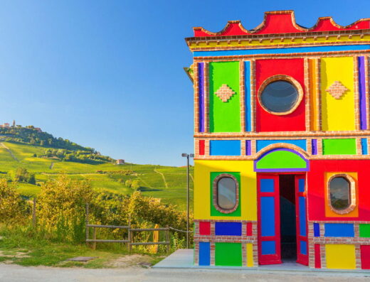 Colorful, two-story building with a decorative facade, reminiscent of a toy house, set against a backdrop of rolling green hills and a distant town.