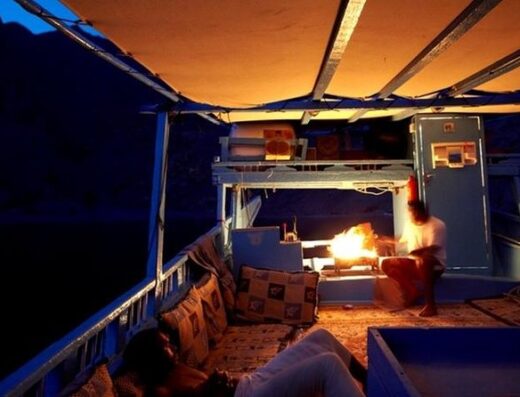 A person sits by a campfire inside a makeshift shelter on a boat, surrounded by mountains at dusk.
