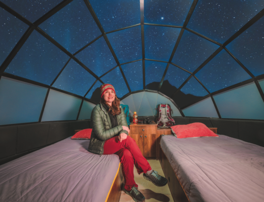 A person sits on a twin bed in a glass-domed room with a starry night sky visible through the ceiling. There are two beds, a small wooden table, and an outdoor backpack in the background.