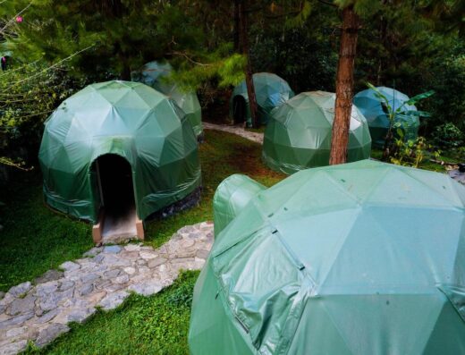 A cluster of green dome-shaped tents arranged in a wooded area, with a stone pathway leading to one of the tents.