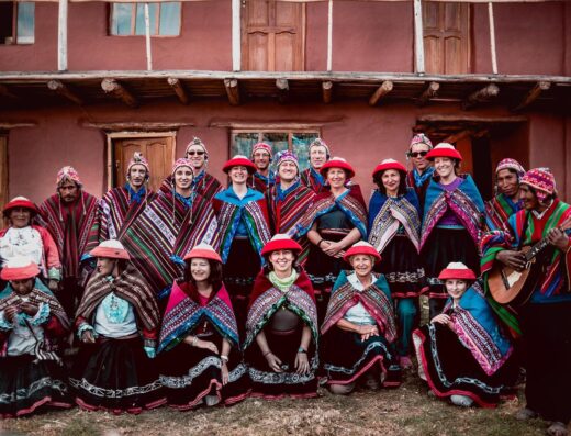 A group of people dressed in traditional clothing stand in front of a building. Some wear brightly colored outfits with patterns and hats, and one person holds a guitar.