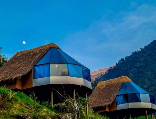 Two modern, triangular huts with thatched roofs situated on a hillside with greenery. A mountain range is in the background and a moon is visible in the early morning or evening sky.