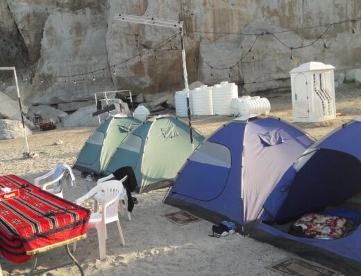 Several tents set up on a sandy beach with limestone cliffs in the background, people nearby, and various camping gear scattered around.
