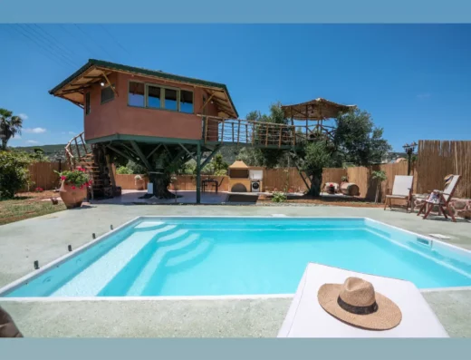 A swimming pool with a lounge chair and hat in the foreground, wooden house elevated on stilts, and clear blue sky in the background.