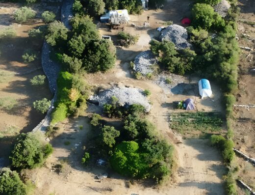 Aerial view of a campsite with several tents, parked vehicles, and camp setups in a wooded area with clearings. Various trees and bushes are dispersed throughout the site.