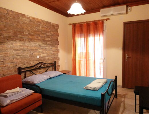A bedroom with a double bed, orange armchair, brick accent wall, TV, air conditioner, and a door with a sheer orange curtain leading to a balcony.