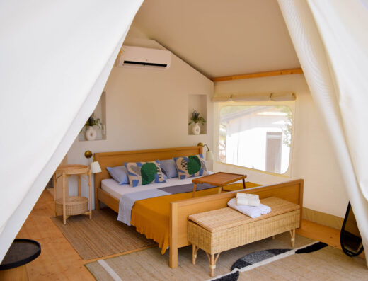 A cozy glamping tent with a neatly made bed, wooden furniture, air conditioning unit, and decorative pillows.