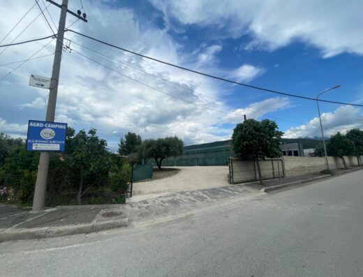 A view of an entrance to an agro-camping site featuring a sign on a utility pole, a gravel driveway, greenery, and a fence under a partly cloudy sky.