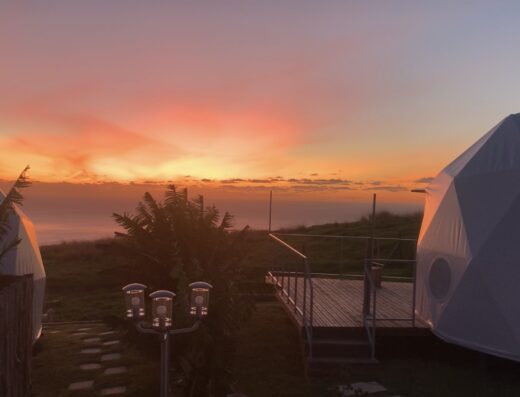 Two white geodesic domes with a wooden deck between them, set against a backdrop of a vibrant sunset over the ocean. Palm trees and a grassy area are in the foreground.