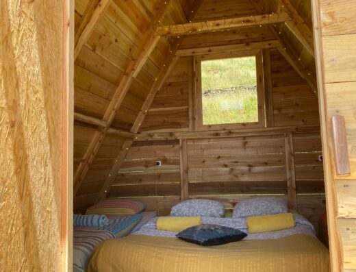 A cozy, wooden cabin interior with a pitched roof, featuring a bed with yellow pillows and blanket, a window above the bed, and a striped rug on the floor.