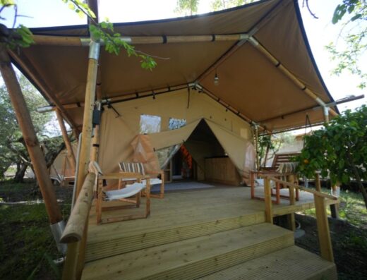 A raised wooden deck with steps leading up to a tan canvas tent structure framed by bamboo poles, featuring outdoor chairs and surrounded by greenery.