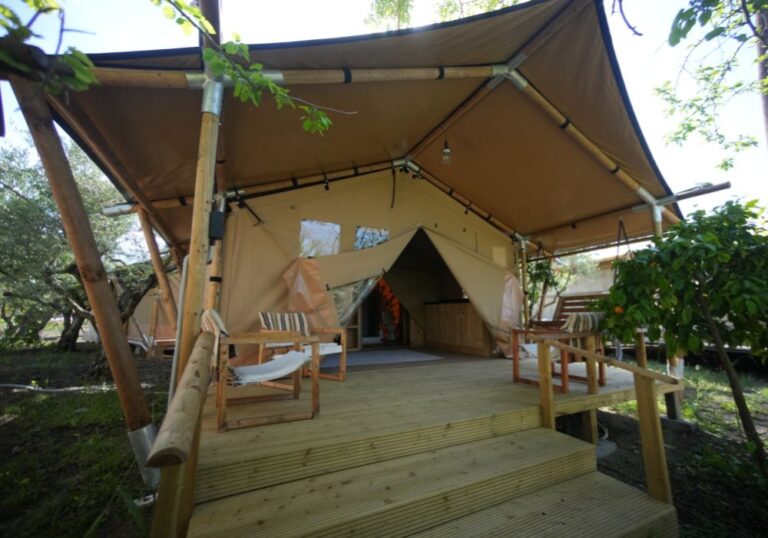 A raised wooden deck with steps leading up to a tan canvas tent structure framed by bamboo poles, featuring outdoor chairs and surrounded by greenery.