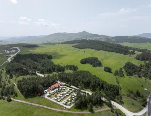 Aerial view of a campsite surrounded by trees and greenery, with rows of white tents or RVs. Fields and hills are visible in the background under a partly cloudy sky.