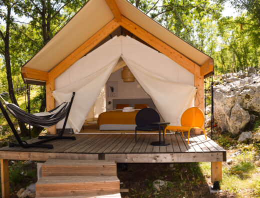 A glamping tent with a small wooden deck featuring a hammock, chairs, and table in a forested area. The tent's door is open, revealing a bed with orange bedding inside.
