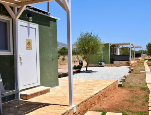 A small green cabin with a white door next to a stone path in an arid area. Another similar cabin and an olive tree are visible in the background.