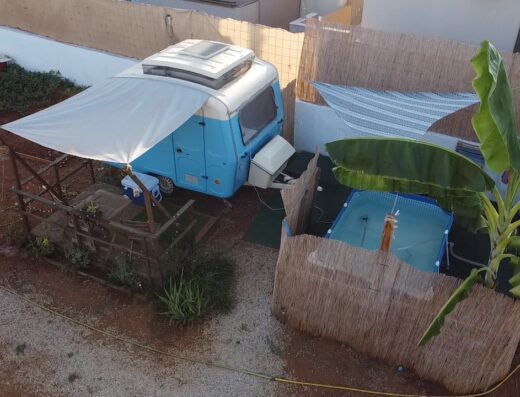Overhead view of a small blue caravan parked beside a makeshift patio with a tented area and a small pool surrounded by bamboo fencing. A banana plant is visible to the right.