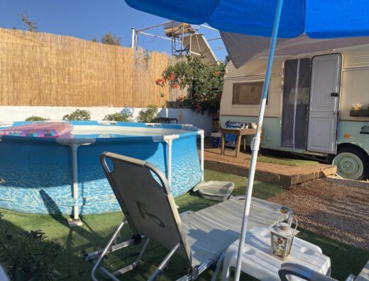 An inflatable swimming pool next to a parked caravan with a wooden fence in the background. Lounge chairs, a blue umbrella, and a lantern are placed on green artificial grass.