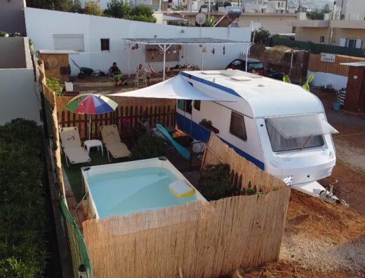 An outdoor setup featuring a parked camper trailer, a pool with two lounge chairs and an umbrella, surrounded by a wooden fence.