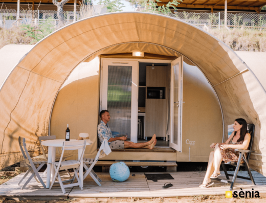 Two people relax outside a small, arched cabin. A man sits on the doorstep with a drink, while a woman relaxes on a chair beside a table with a wine bottle and glasses.