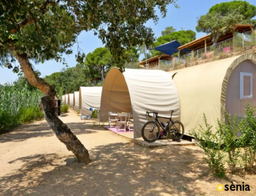 A row of beige canvas tents with small porches and a bicycle parked in front. Surrounded by trees and greenery, under a clear blue sky.