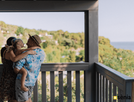 Three people, one with a straw hat, are standing on a balcony overlooking a lush landscape and ocean.