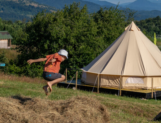 A person in a hat and shorts is jumping onto a pile of hay near a large beige tent, with green trees and hills in the background.