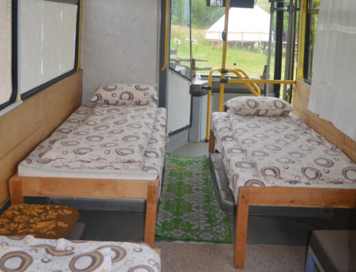 Interior of a converted bus with two single beds, patterned bedspreads, a green rug, and a small table, with windows letting in natural light.