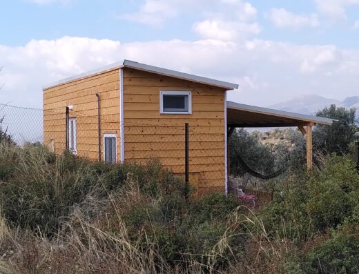 A small, modern wooden cabin with a sloped roof and covered porch stands among dry grass and shrubs in a rural landscape.